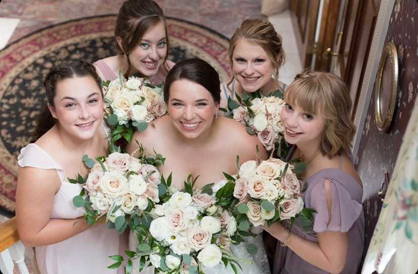 Bride and bridesmaids with bouquets of white and blush roses, smiling and posing together.