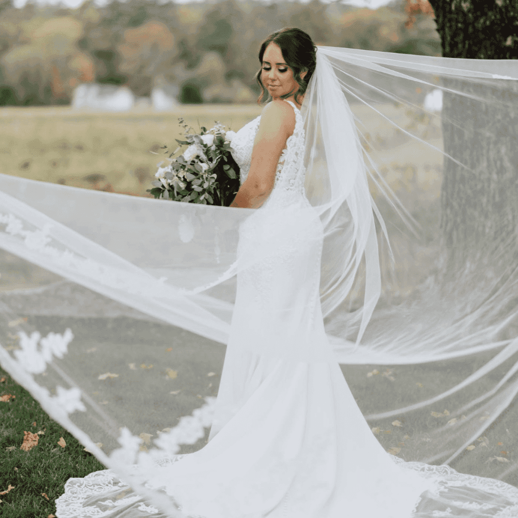 Bride in white gown holding bouquet, with veil flowing in outdoor setting.