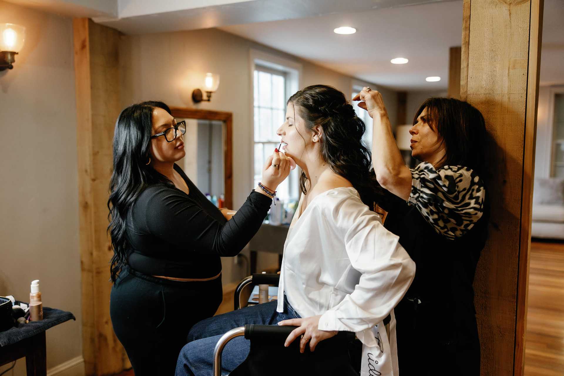 Bridesmaids help each other with hair and makeup in a cozy room.