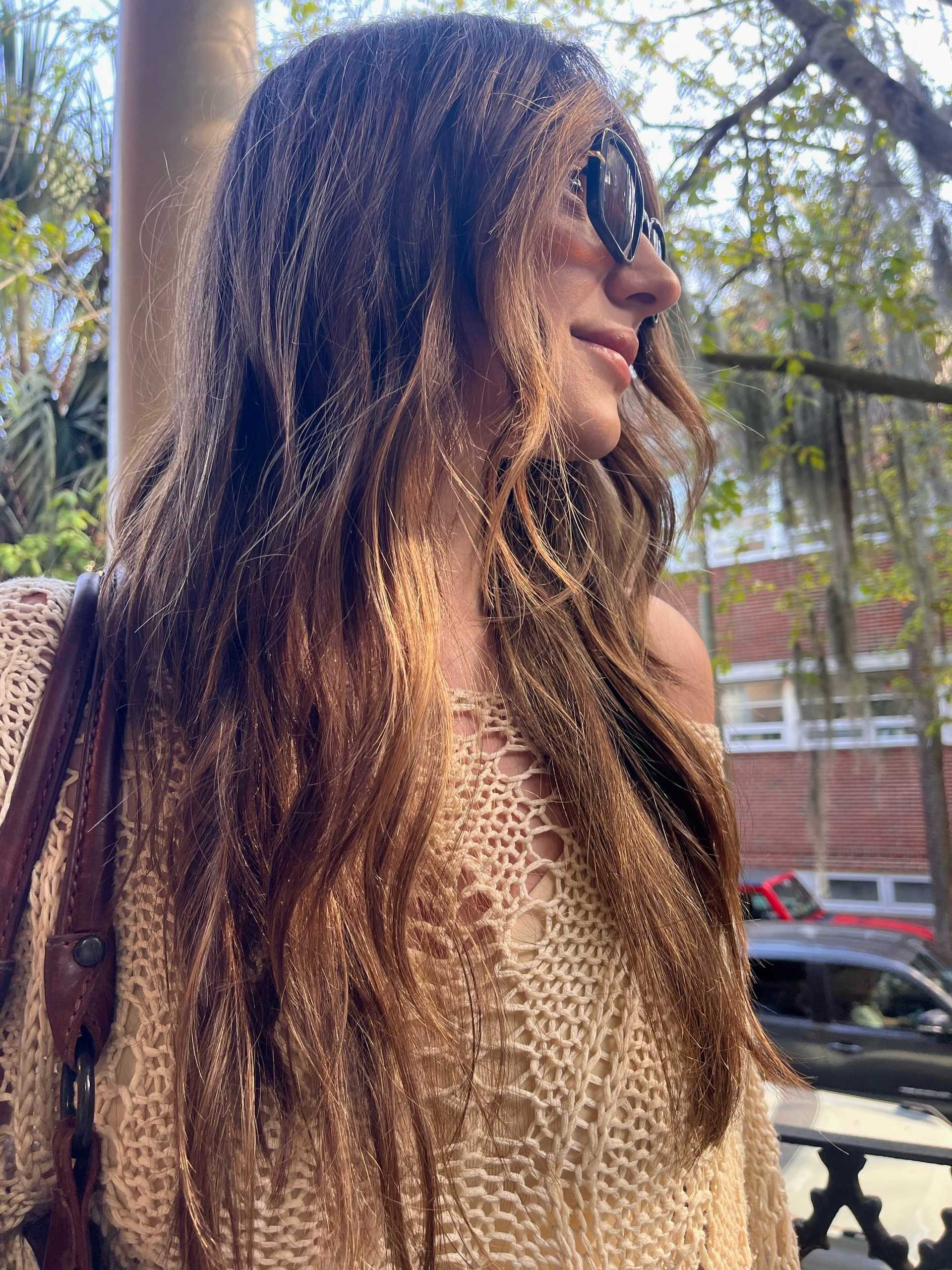 Woman in sunglasses smiling outdoors, surrounded by greenery and urban elements.