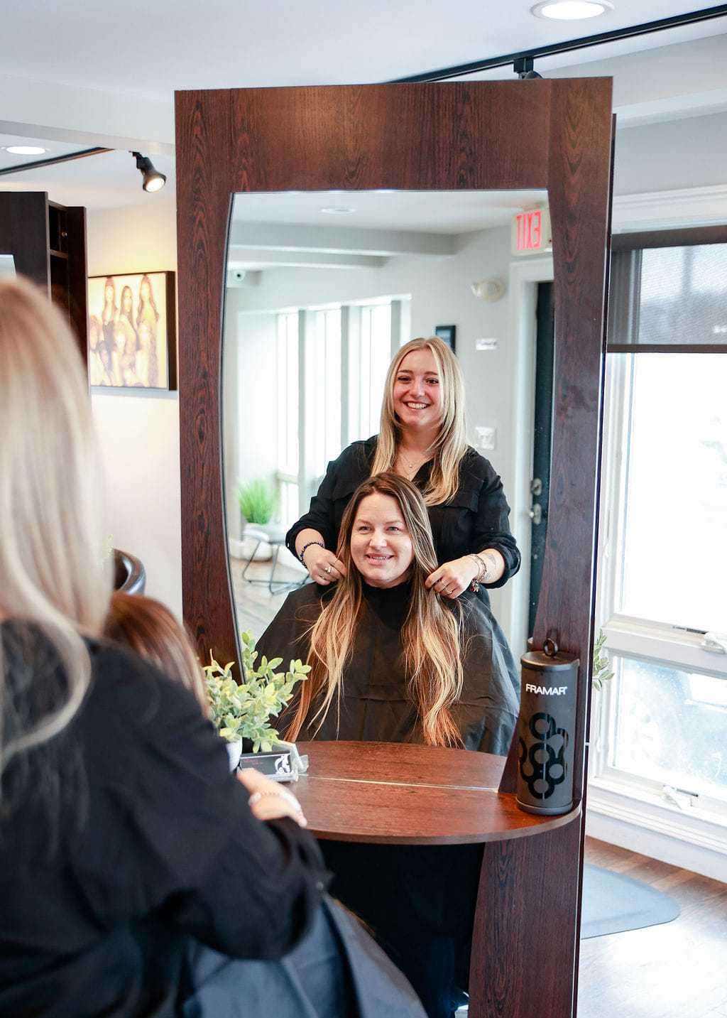Hairdresser styling long hair in a salon, with client smiling in the mirror.