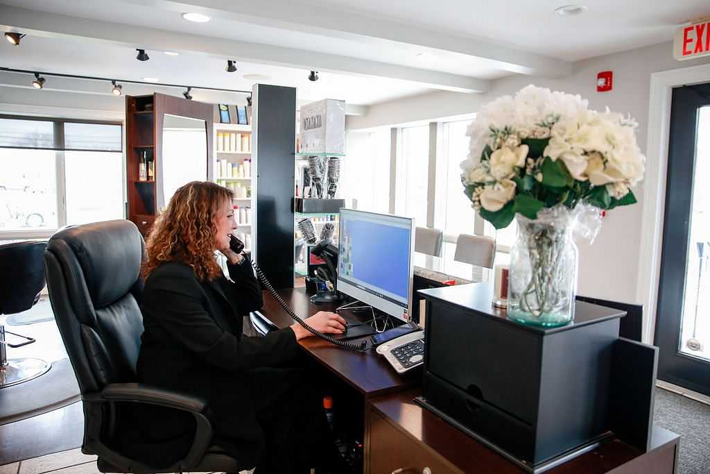 Receptionist at desk in modern office, using phone and computer, white flowers on counter.