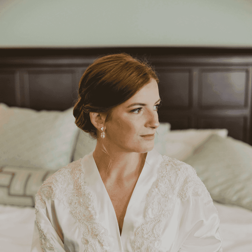 Bride in embroidered robe, sitting on bed, looking sideways pensively before wedding ceremony.